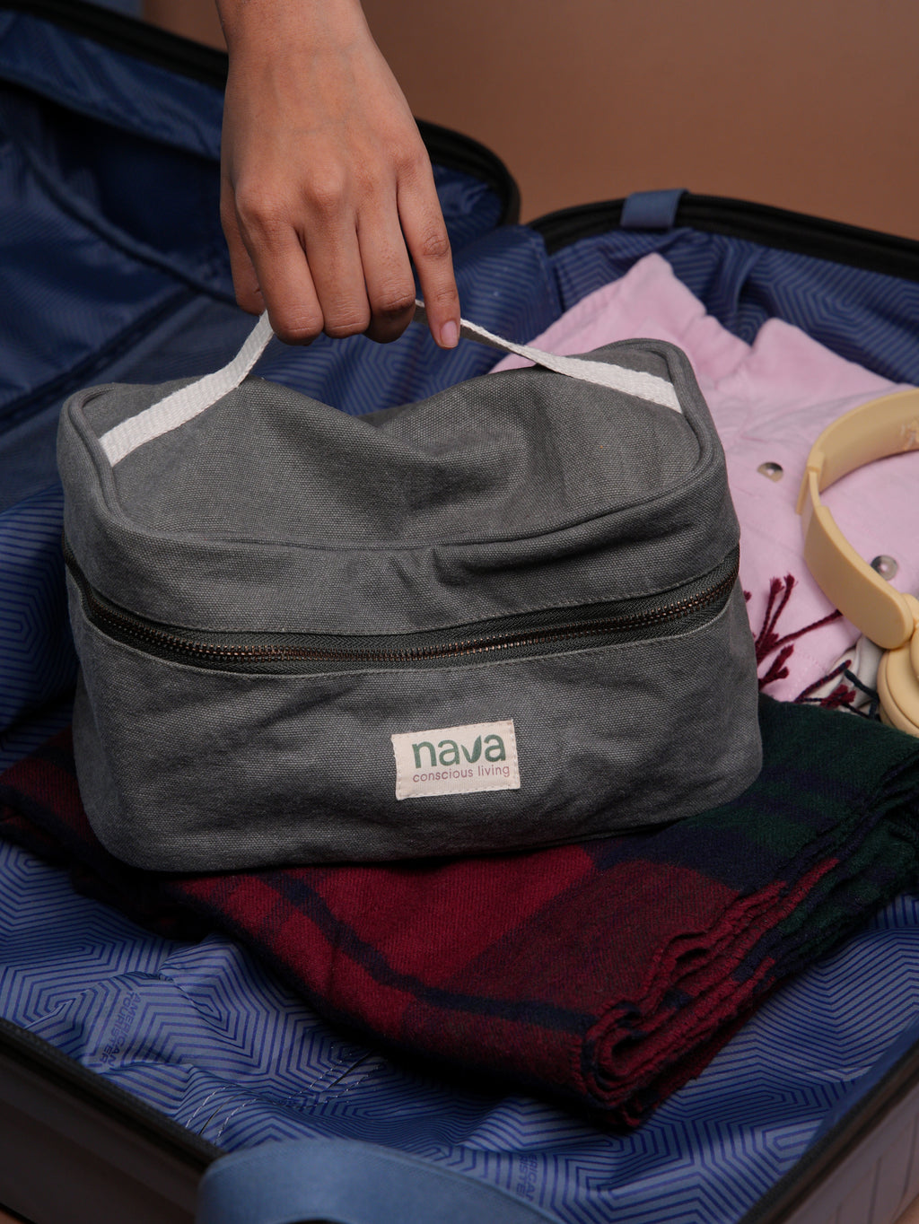 Gray toiletry bag with 'nava' brand label being placed into a suitcase filled with clothes.